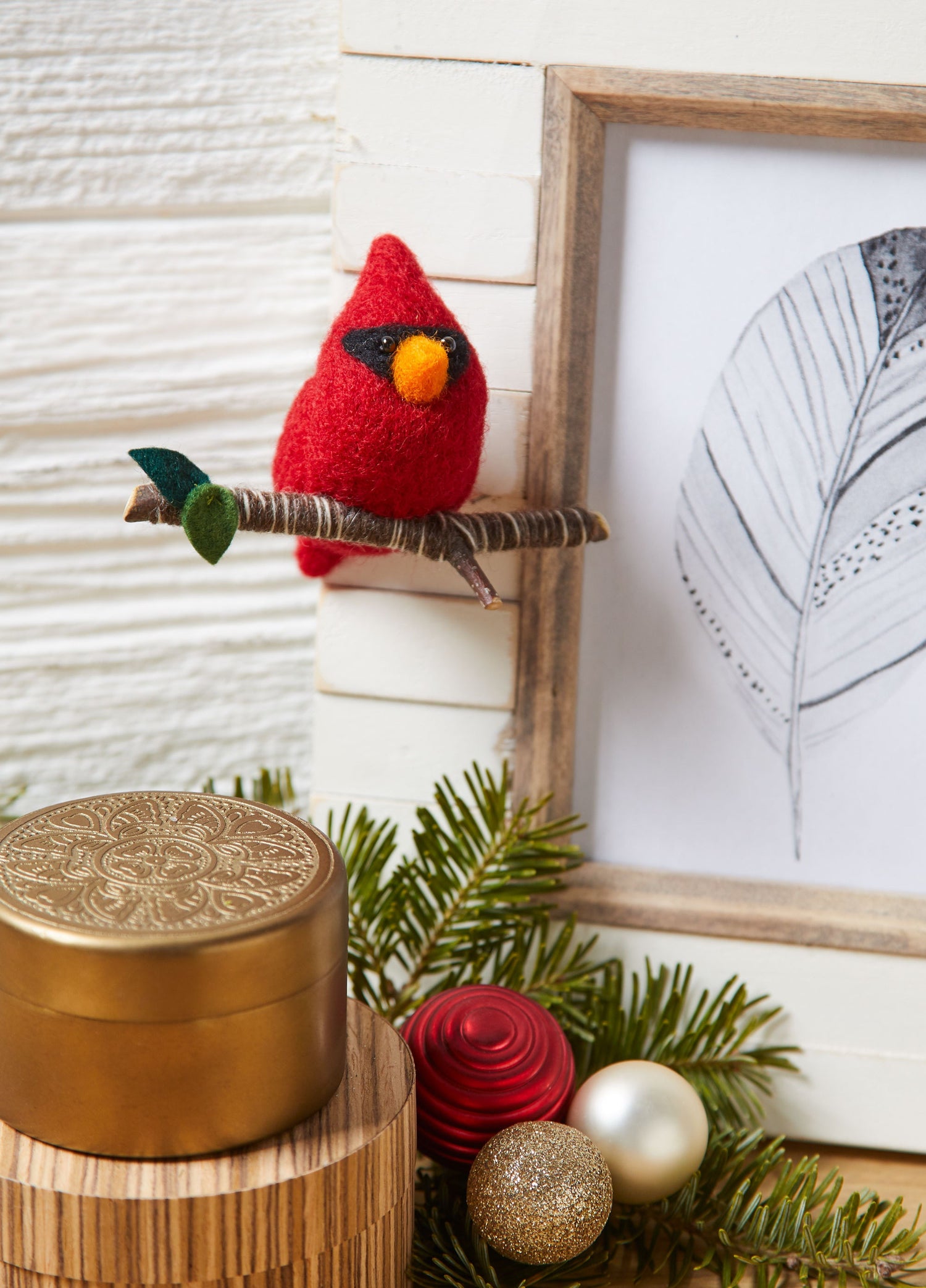 Holiday decor scene with white wood background, two golden tins on table, picture frame with pencil-drawn leaf art, and a needle-felted three-inch cardinal bird ornament