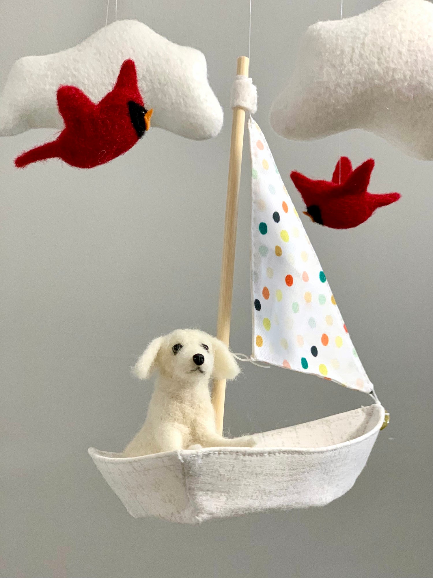 Mobile of a small white needle-felted dog sitting in a sailboat with two red cardinals flying above along with two felt clouds.
