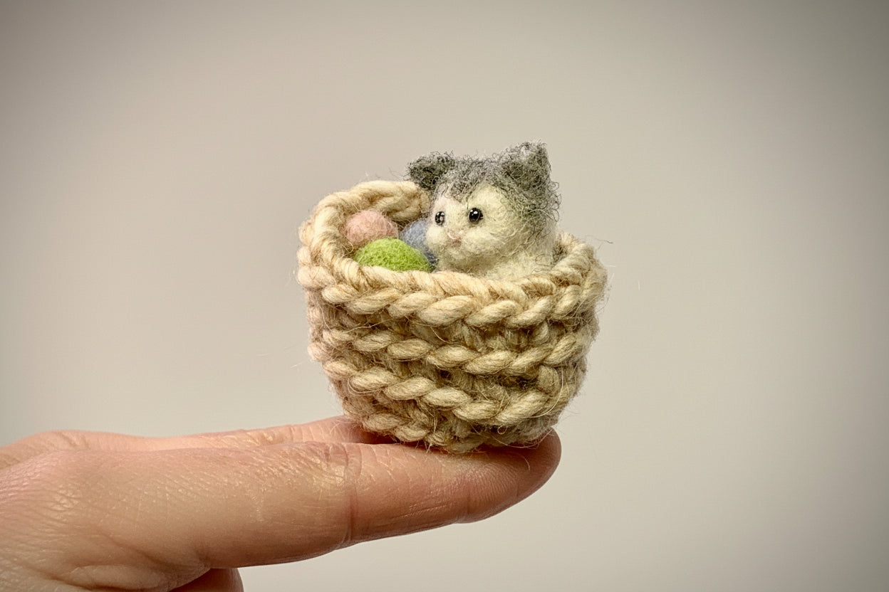 Small knitted basket with a needle felted  cat surrounded by yarn held by a hand against a neutral background
