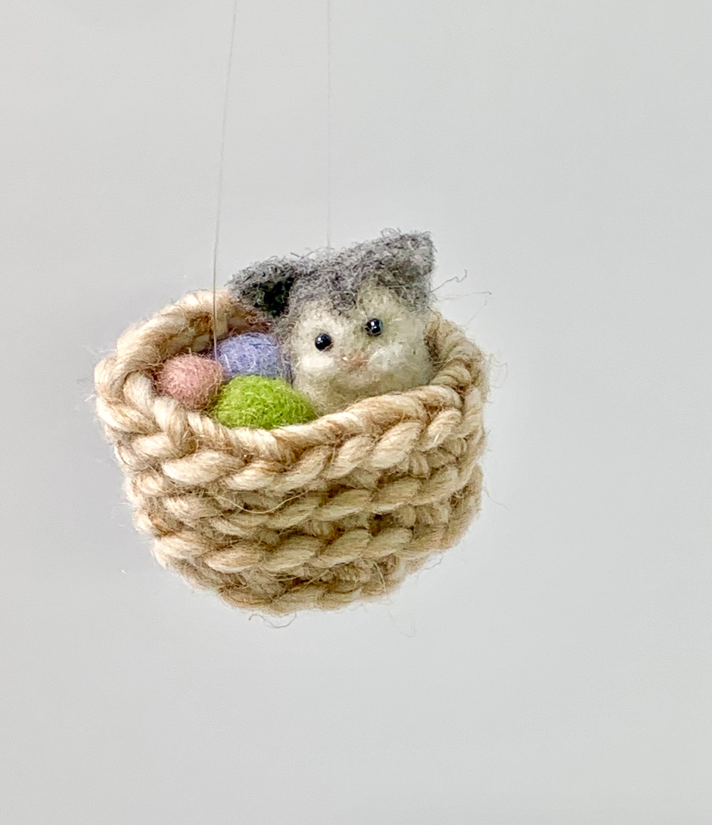 Small needle felted gray and white kitten sitting in a woven basket  surround by yarn on a light gray background
