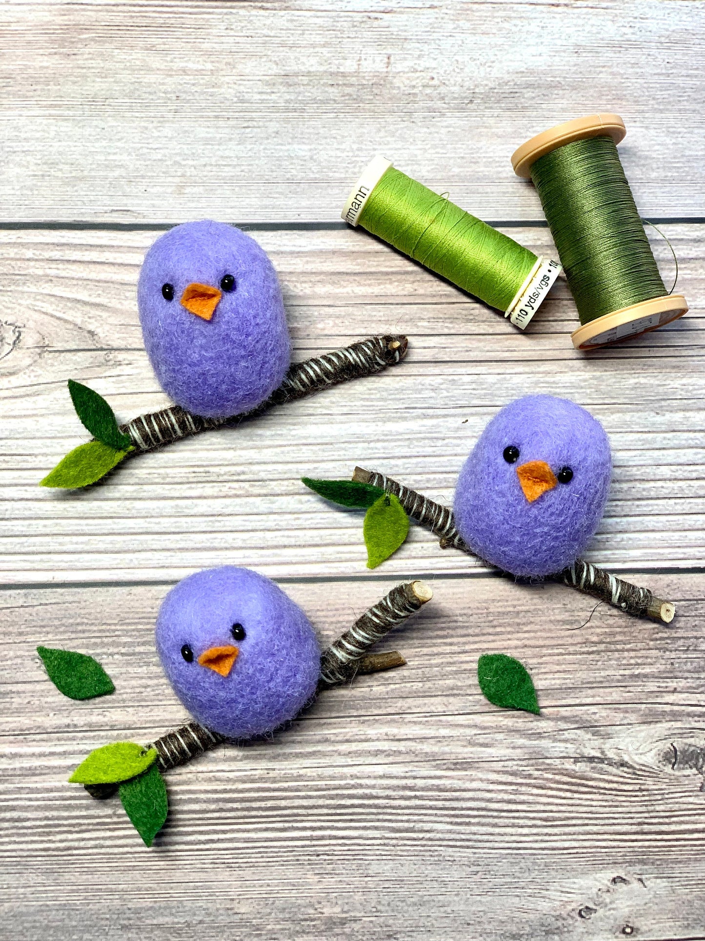Three needle-felted wool purple birds on branches placed on wooden table